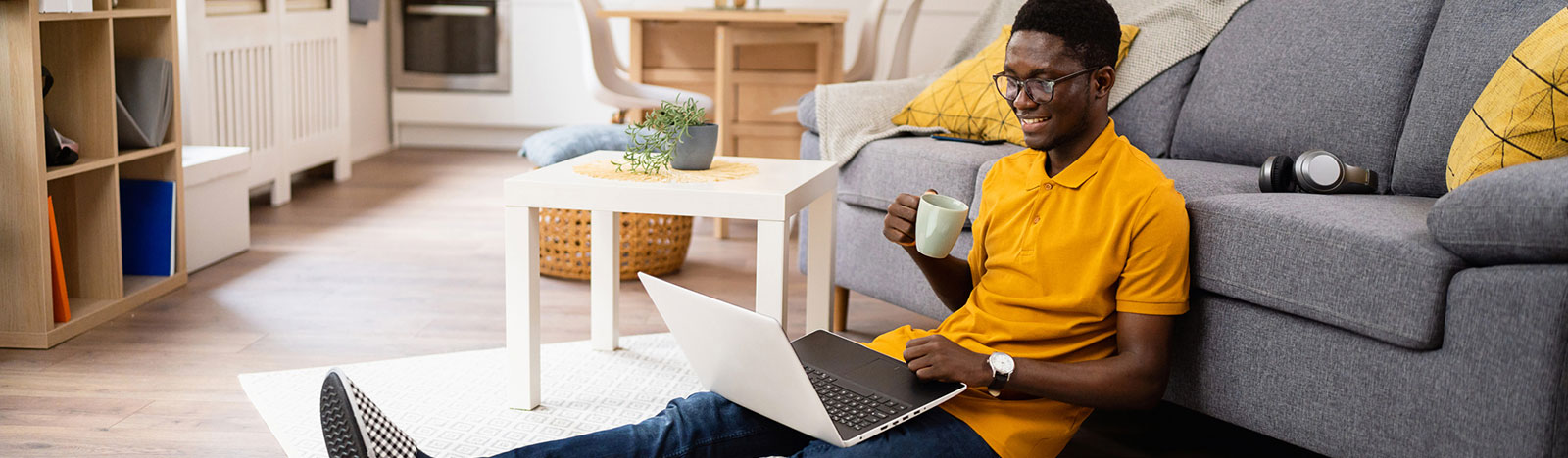 Modern and young African-American male freelancer, drinking coffee and working on laptop while sitting on the floor in the living room