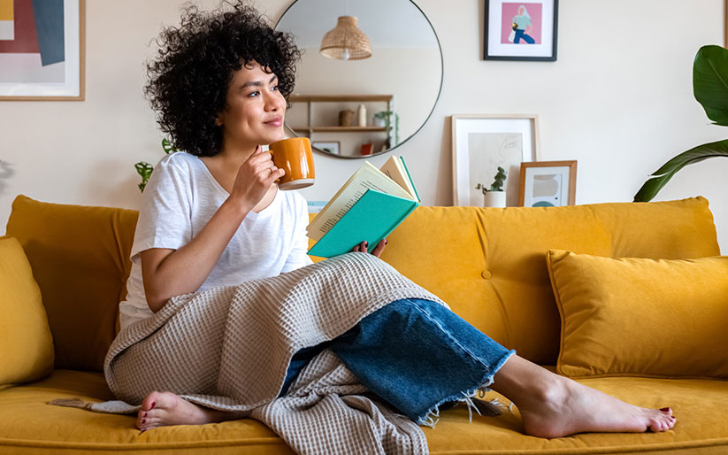 African american woman reading a book at home, drinking coffee sitting on the couch.